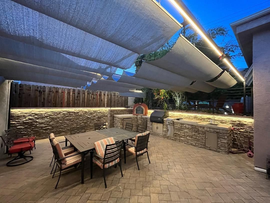 Modern outdoor patio with shaded seating, dining area, stone walls, and BBQ grill setup.